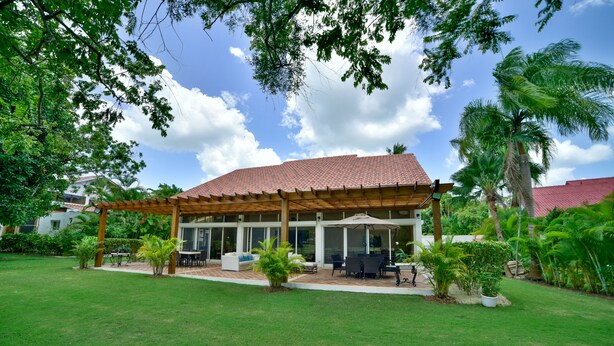 A covered outdoor seating area with tables and chairs, surrounded by lush greenery and a clear sky. - casa de campo villa 204 (La Romana)