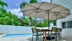 A poolside area with a table and chairs under a large umbrella, surrounded by lush greenery and a white wall. - casa de campo villa 204 (La Romana)