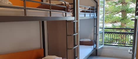 A dormitory room with bunk beds, a wooden floor, and a balcony with a view of trees.
