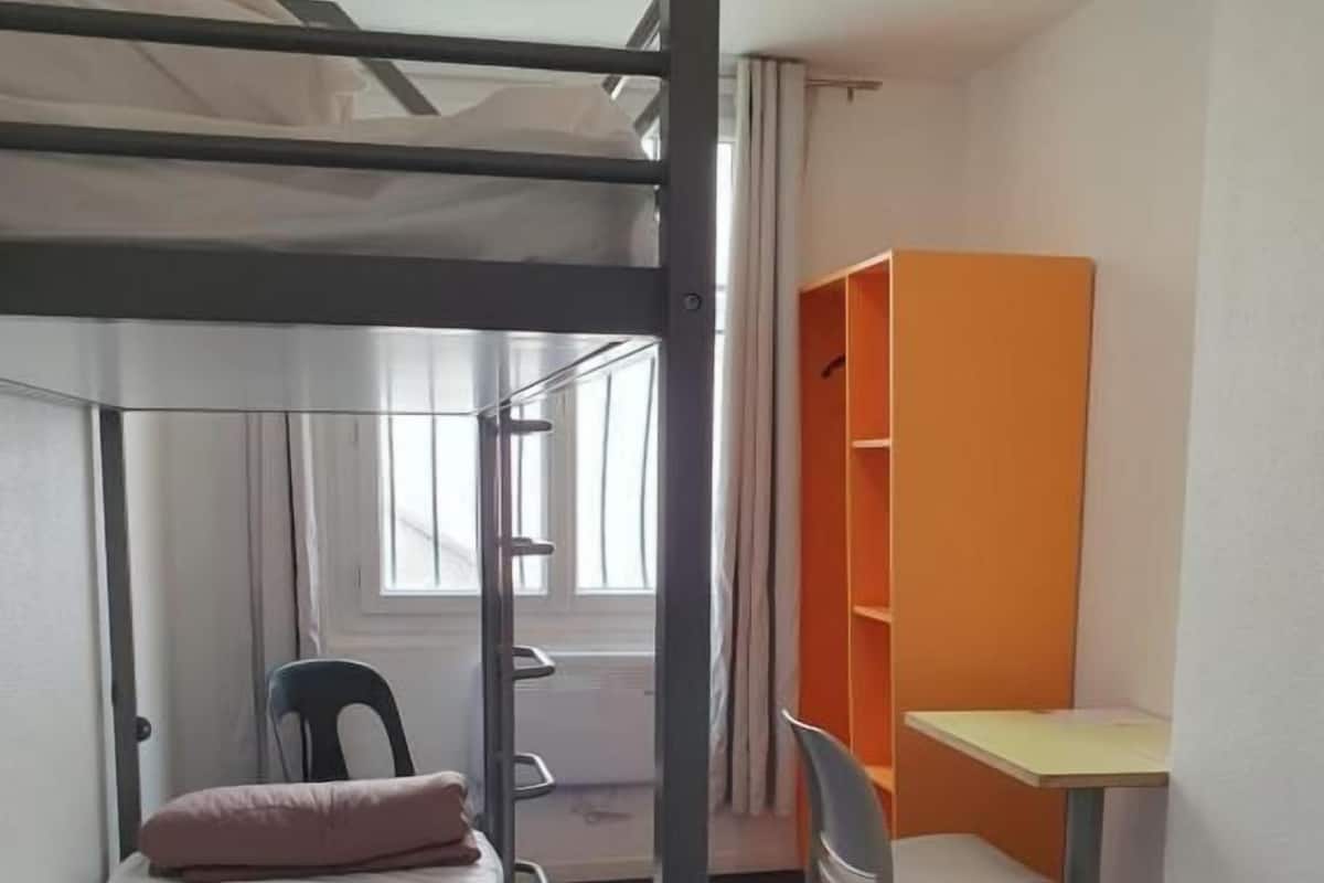 A bunk bed with a desk and chair, a window, and an orange shelving unit in a room.