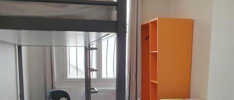 Uma cama beliche com escrivaninha e cadeira, uma janela e uma estante laranja num quarto.