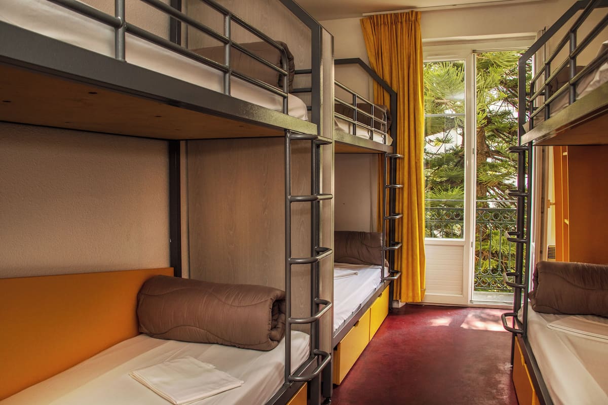 A dormitory-style room with bunk beds, a window with yellow curtains, and a view of greenery outside.