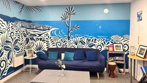 A living room with a large mural of a coastal scene, a dark blue sofa with teal cushions, a small coffee table with a vase, and a bookshelf with framed photos and books. - Auberge de Jeunesse HI Nice - Les Camélias (Nice)