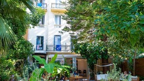 An outdoor café area with tables and chairs, surrounded by lush greenery and a building with balconies. - Auberge de Jeunesse HI Nice - Les Camélias (Nice)