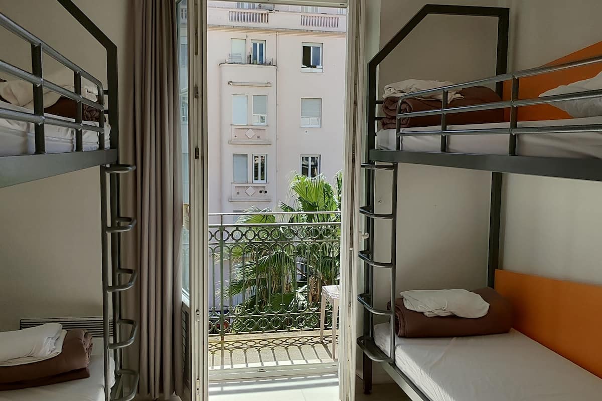 A dormitory room with bunk beds, a balcony with a metal railing, and a view of a building with multiple windows.