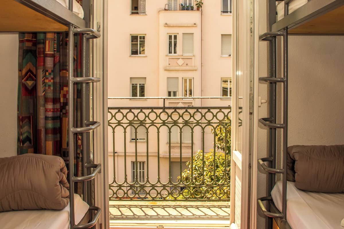 A narrow corridor with bunk beds on either side, leading to an open balcony with a view of a building with balconies.