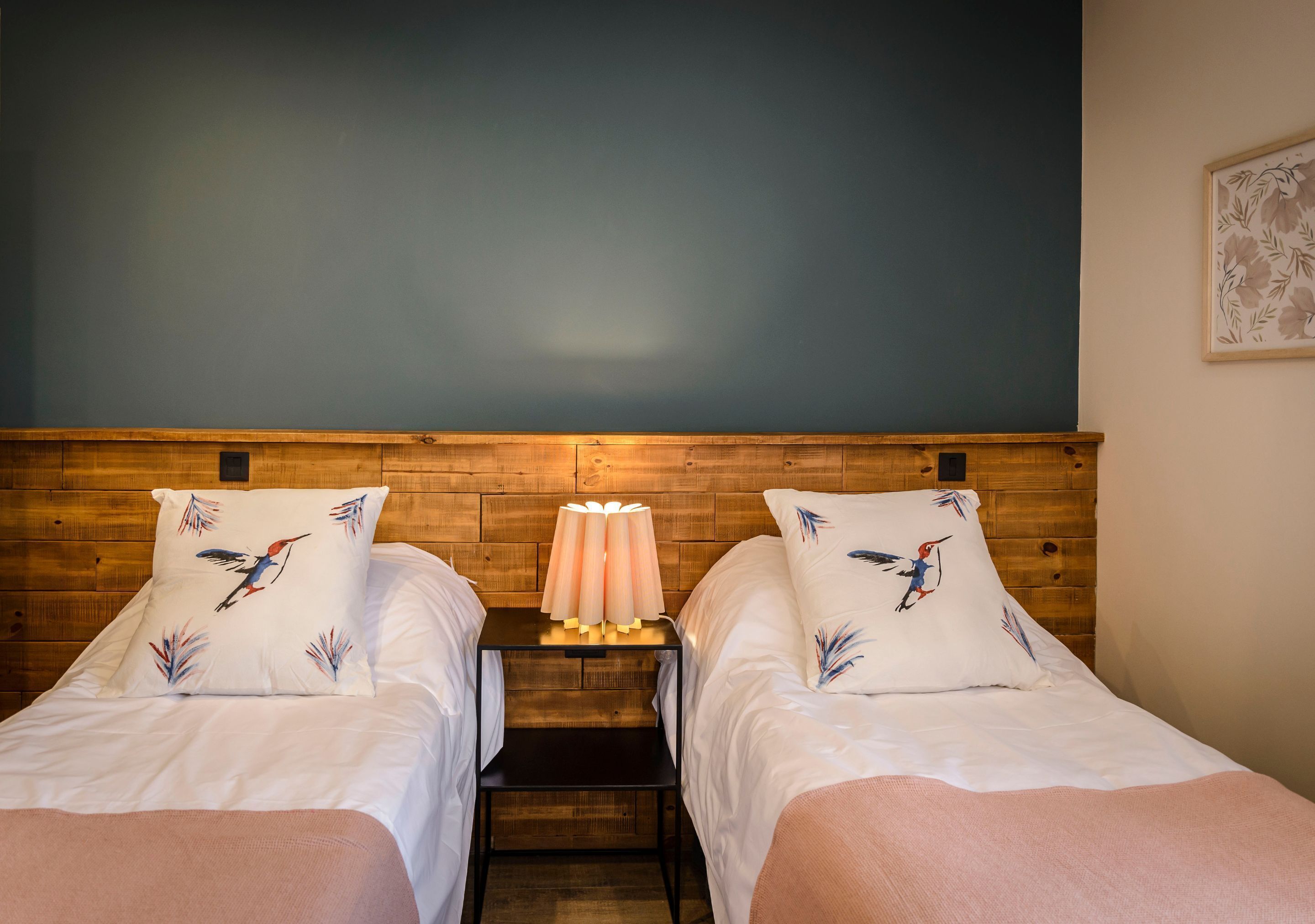 A room with two beds, a wooden headboard, a nightstand with a lamp, and a floral wall decoration.