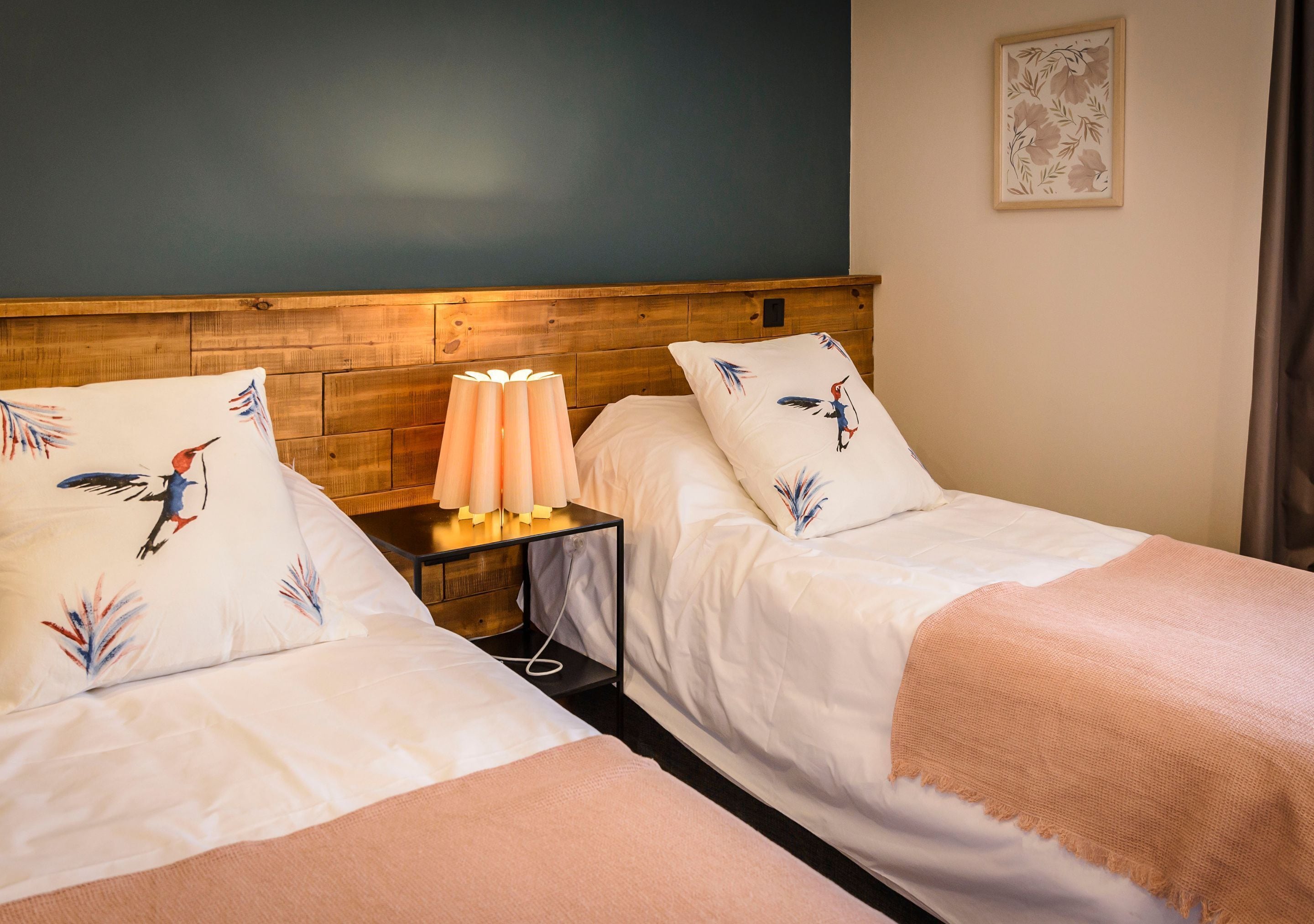 Two single beds with matching headboards and pillows, a bedside table with a lamp, and a framed floral artwork on the wall.