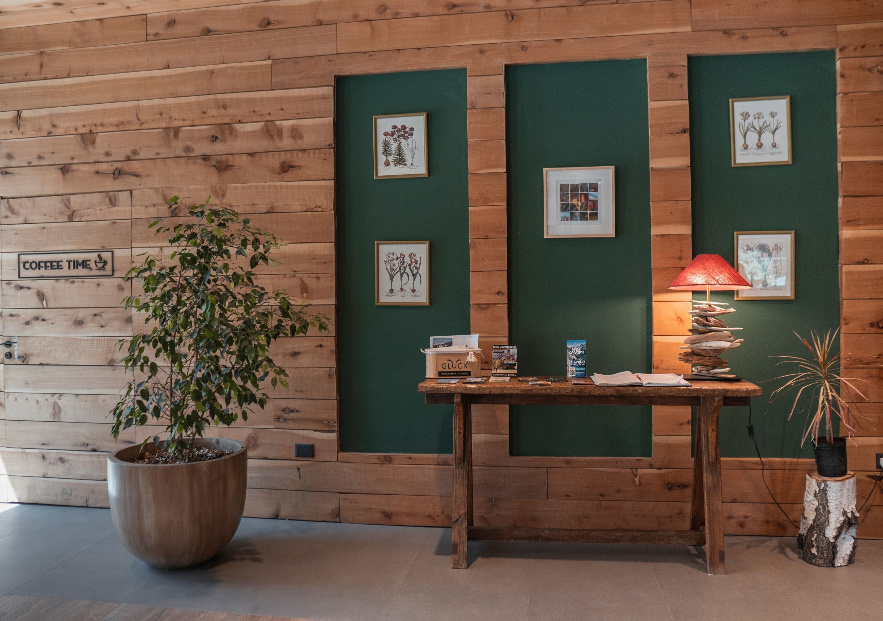 A wooden wall with framed botanical illustrations, a coffee sign, and a table with a lamp and books.