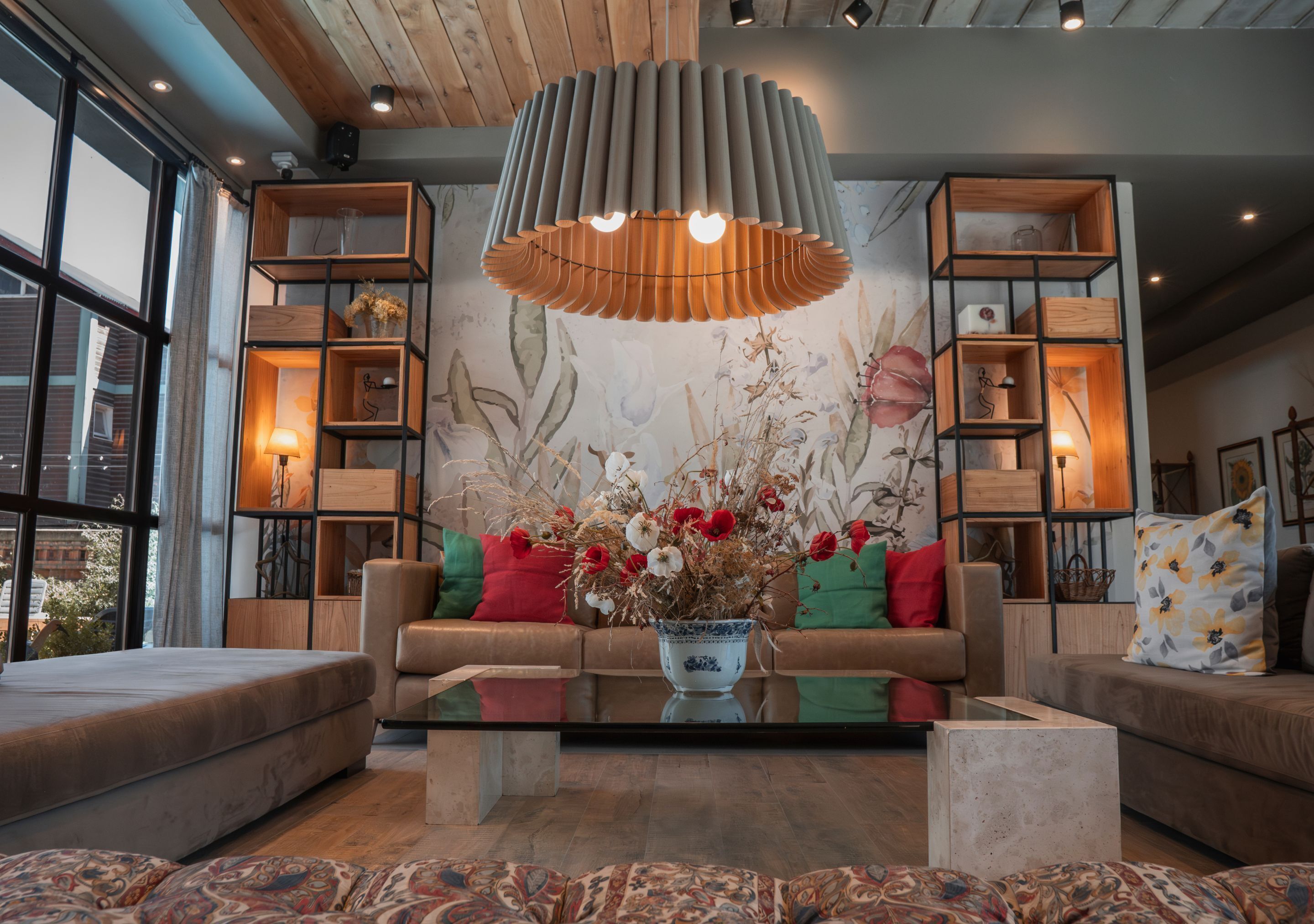 A modern living room with a sofa set, a coffee table, and a decorative vase with flowers. There are wooden shelves on either side of the sofa, and a large window on the left side.