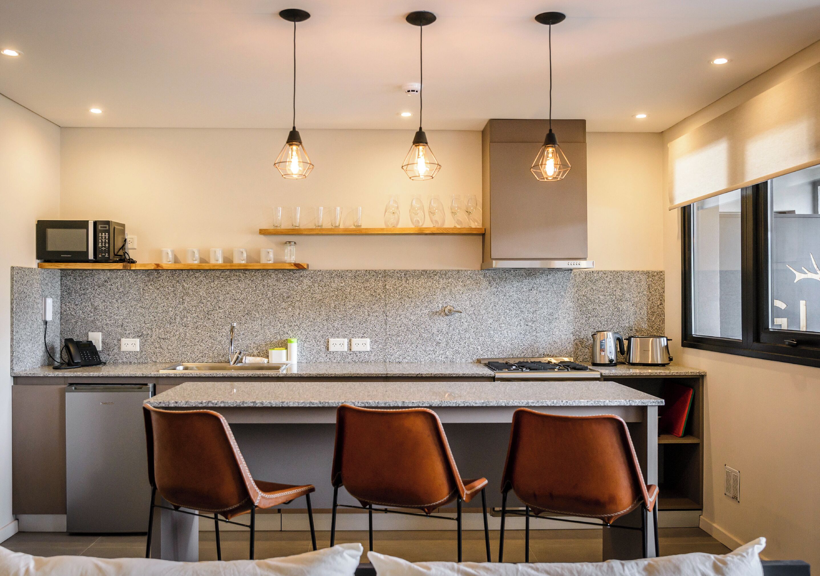 Une cuisine moderne dotée de plans de travail en granit, d’un îlot central avec des tabourets de bar, d’un éclairage suspendu et d’appareils électroménagers intégrés.