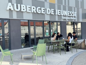 Ett utomhusområde med bord och stolar framför en byggnad där det står ett skylt med texten ”AUBERGE DE JEUNESSE Jacques Delors”.