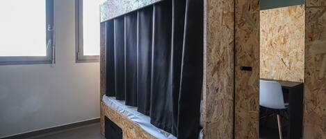 A bunk bed with a wooden headboard and black curtains, a small desk with a chair, and a window providing natural light.