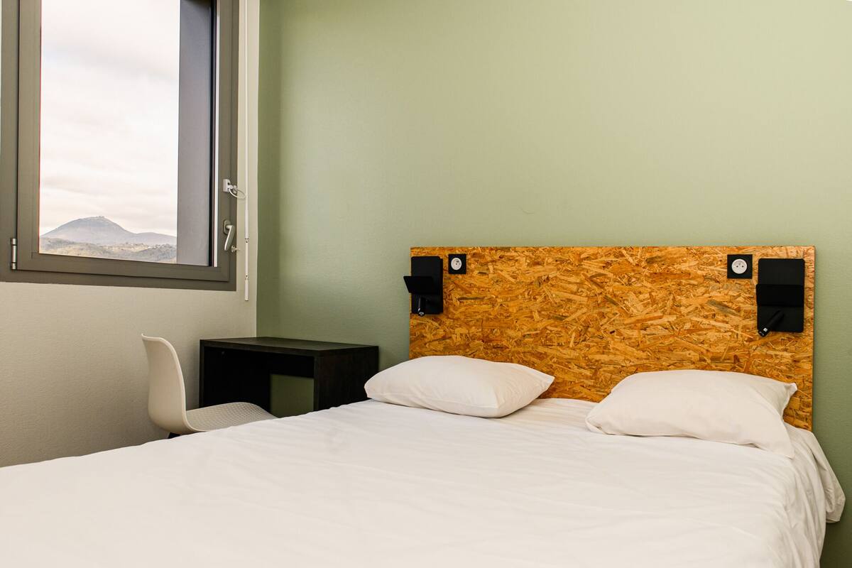 A bedroom with a bed, a chair, a nightstand, and a window with a view of a mountain.
