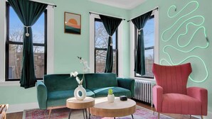 Un salon avec un canapé de couleur turquoise, un fauteuil rose et une table basse ronde. Il y a deux fenêtres avec des rideaux foncés, un radiateur et un néon fixé au mur.