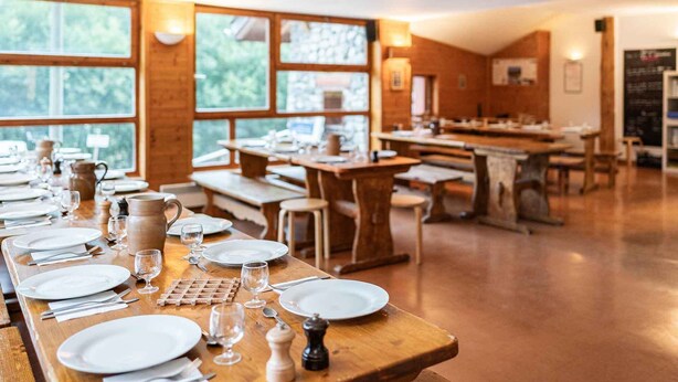 Een restaurant met houten tafels en banken, grote ramen en een menubord.