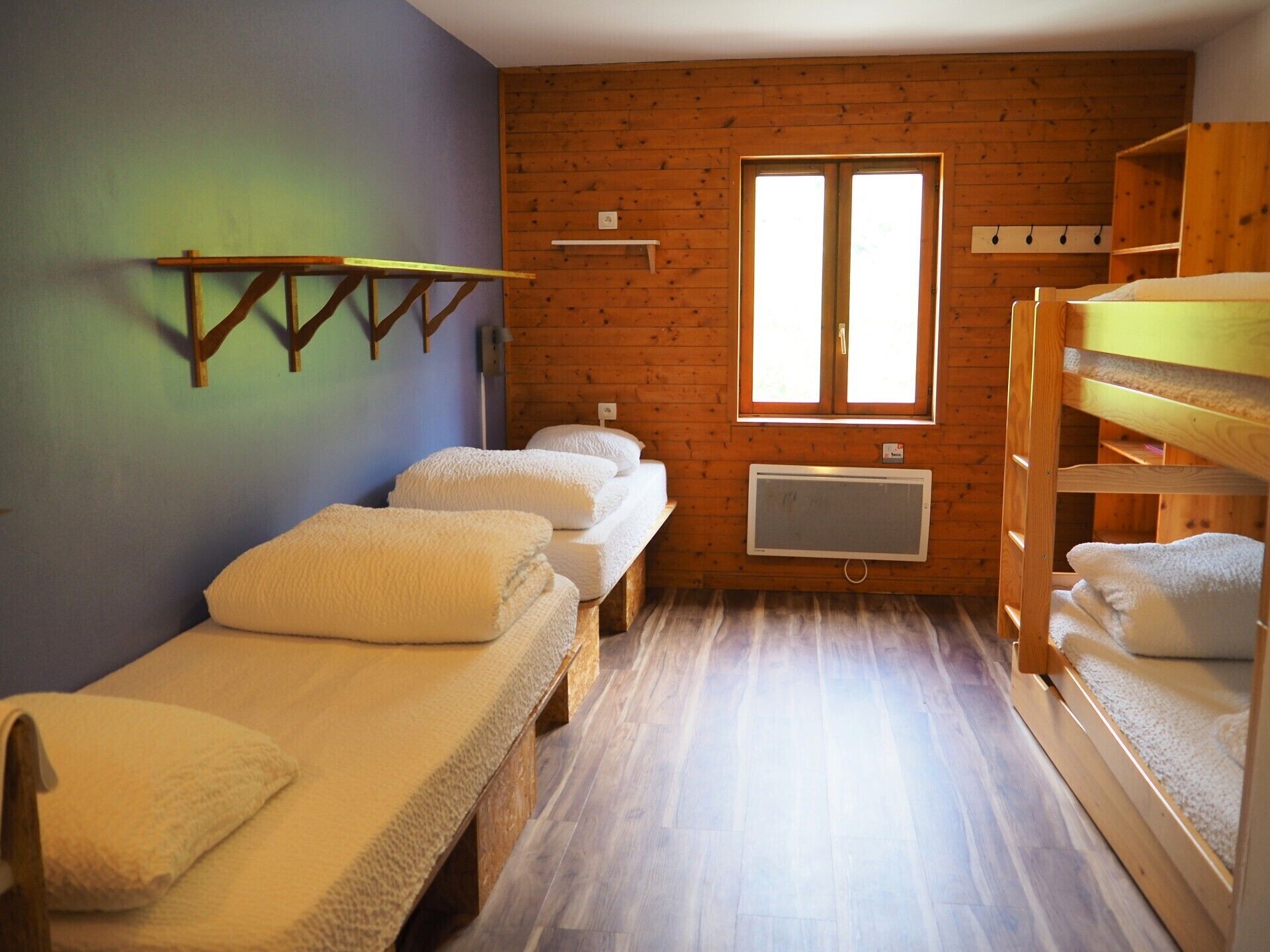 Een kleine, knusse kamer met twee eenpersoonsbedden, een houten stapelbed, een raam met daglicht en een wandradiator.