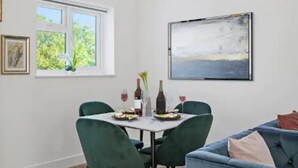Un comedor con una mesa preparada para dos personas, dos sillas, un ventanal con vistas a la vegetación y un cuadro en la pared.