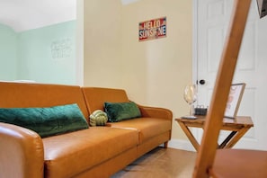 A living room with a brown leather sofa, green cushions, a small wooden side table with a glass, and a 'Hello Sunshine' sign on the wall.