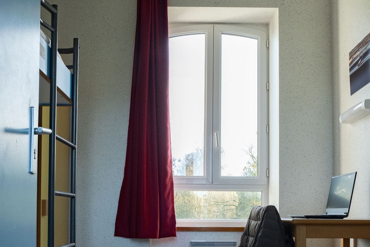 Un dormitorio con literas, un escritorio con una computadora portátil, una ventana con cortina y un radiador.