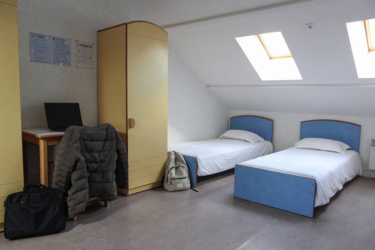Habitación tipo dormitorio con dos camas individuales, un escritorio con una computadora portátil, un armario y un tragaluz.