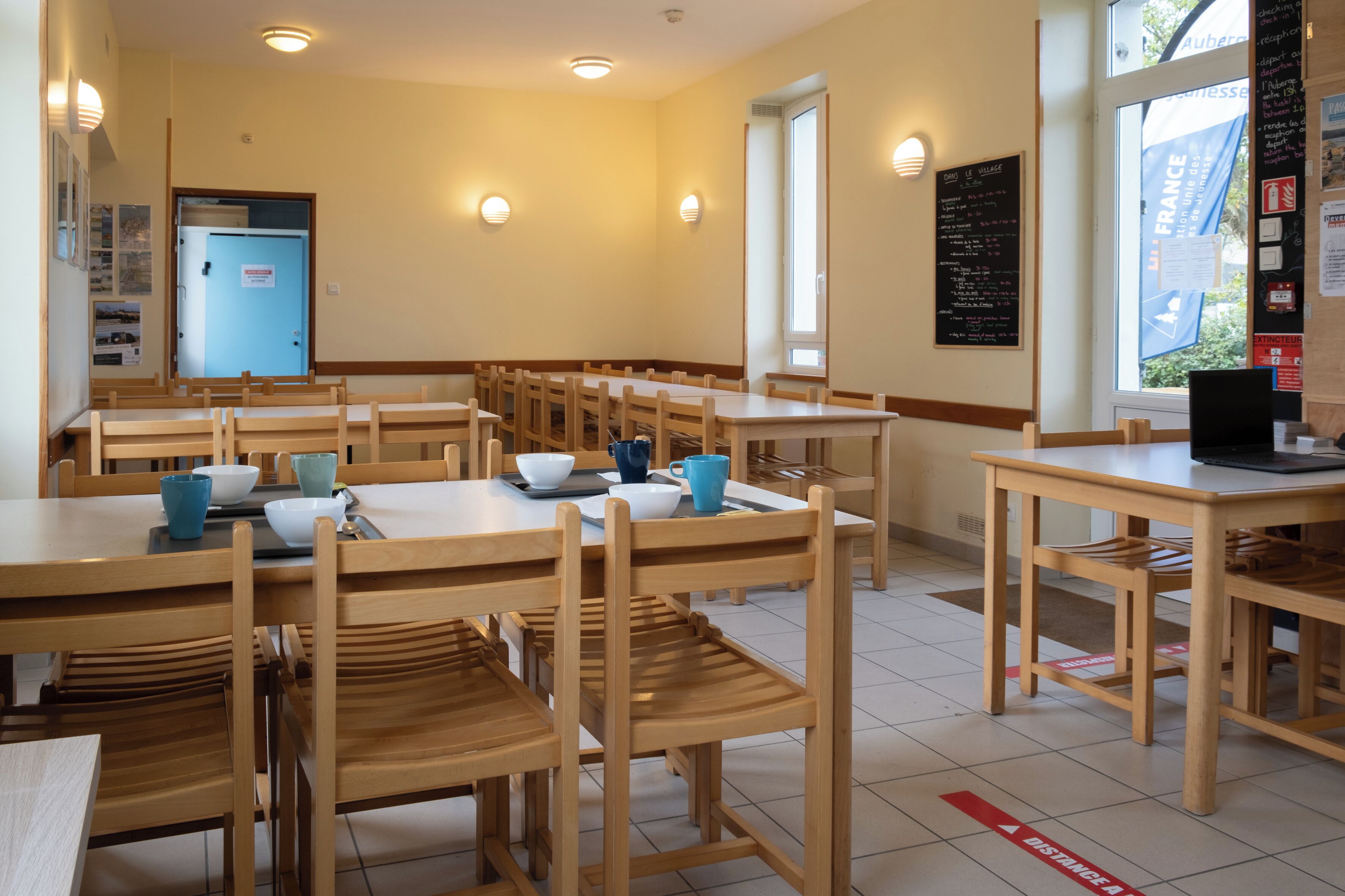 A dining area with multiple wooden tables and chairs, a blue door, and a window with a view of the outside.