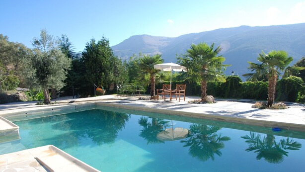 Piscina cristalina con vista a montañas al fondo, rodeada de palmeras y un área de patio con sillas y una sombrilla.