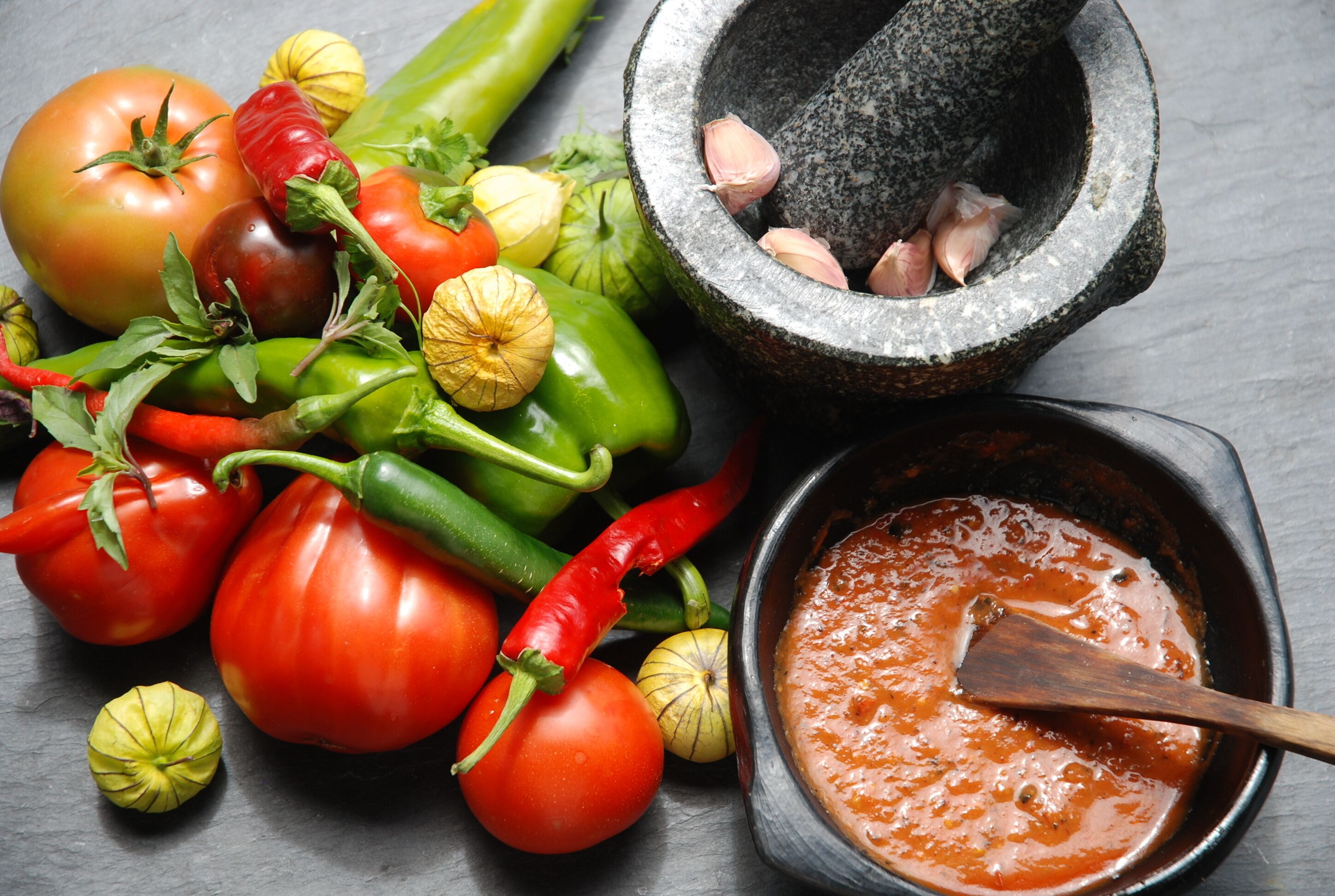 A variety of fresh vegetables including tomatoes, peppers, and garlic, along with a mortar and pestle and a bowl of sauce.
