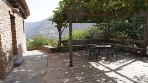 A wooden deck with stone flooring, a pergola with grapevines, and a view of mountains.