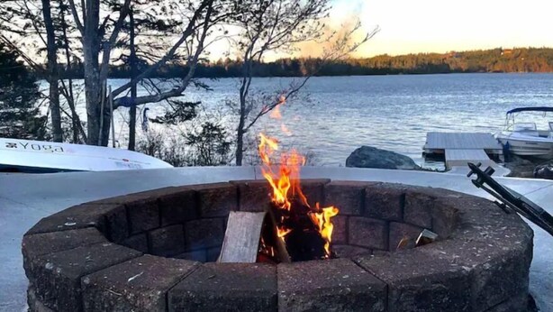 Un pozo de fuego de piedra circular con llamas, rodeado por una masa de agua y barcos al fondo.