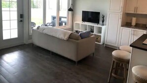 Una sala de estar moderna con un sofá, un televisor sobre un mueble bajo y una zona de cocina con armarios y un taburete de bar.