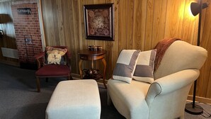 A cozy room with a wooden paneled wall, a framed map, a red armchair, a cream-colored armchair, and a small table with candles.