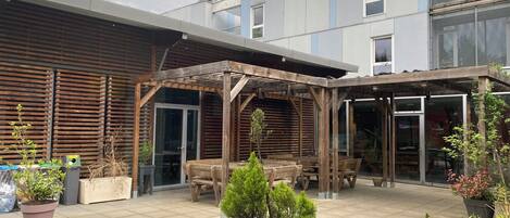 Un patio cubierto con mobiliario de madera, plantas en macetas y un edificio de diseño moderno al fondo.