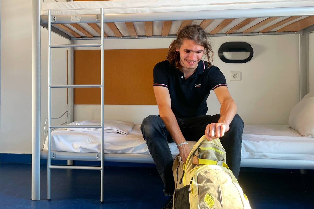 A bunk bed room with a metal ladder, a white mattress on the lower bunk, and a person sitting on the floor with a backpack.