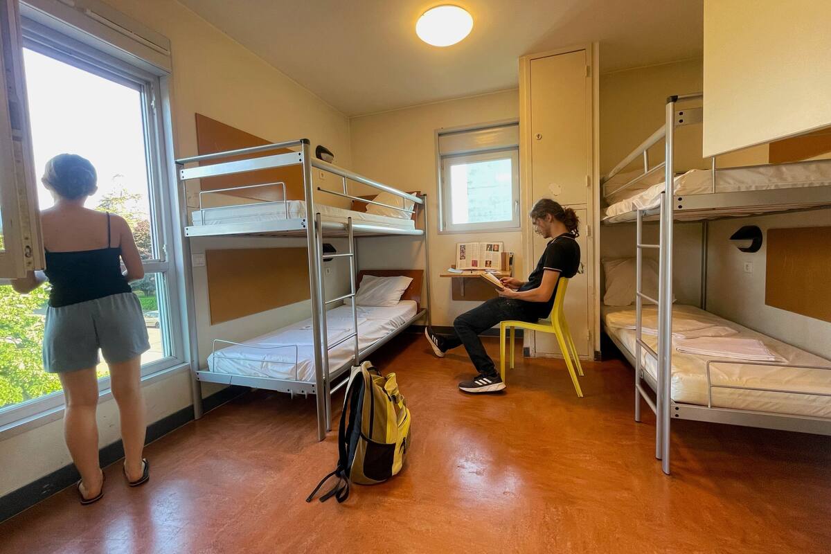 A dormitory room with bunk beds, a window, and a desk with books.