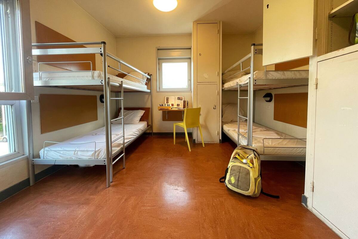 A dormitory room with bunk beds, a desk, a chair, and a backpack.