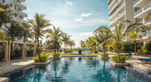 A swimming pool surrounded by palm trees and lounge chairs, with modern buildings in the background. - 2ME-4 APARTAMENTO DE 2 ALC FRENTE AL MAR (Cartagena)