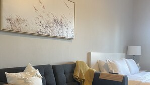 A modern hotel room with a grey sofa, a bed with white bedding, and a large abstract painting on the wall. - Apartment 202 (Cando)
