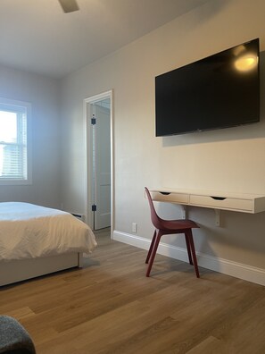 A bedroom with a bed, a desk, a chair, a TV mounted on the wall, and a door leading to another room. - Apartment 202 (Cando)
