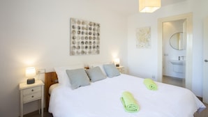 Un dormitorio con una cama grande, dos mesitas de noche con lámparas, una obra de arte en la pared y un baño visible a través de una puerta abierta.