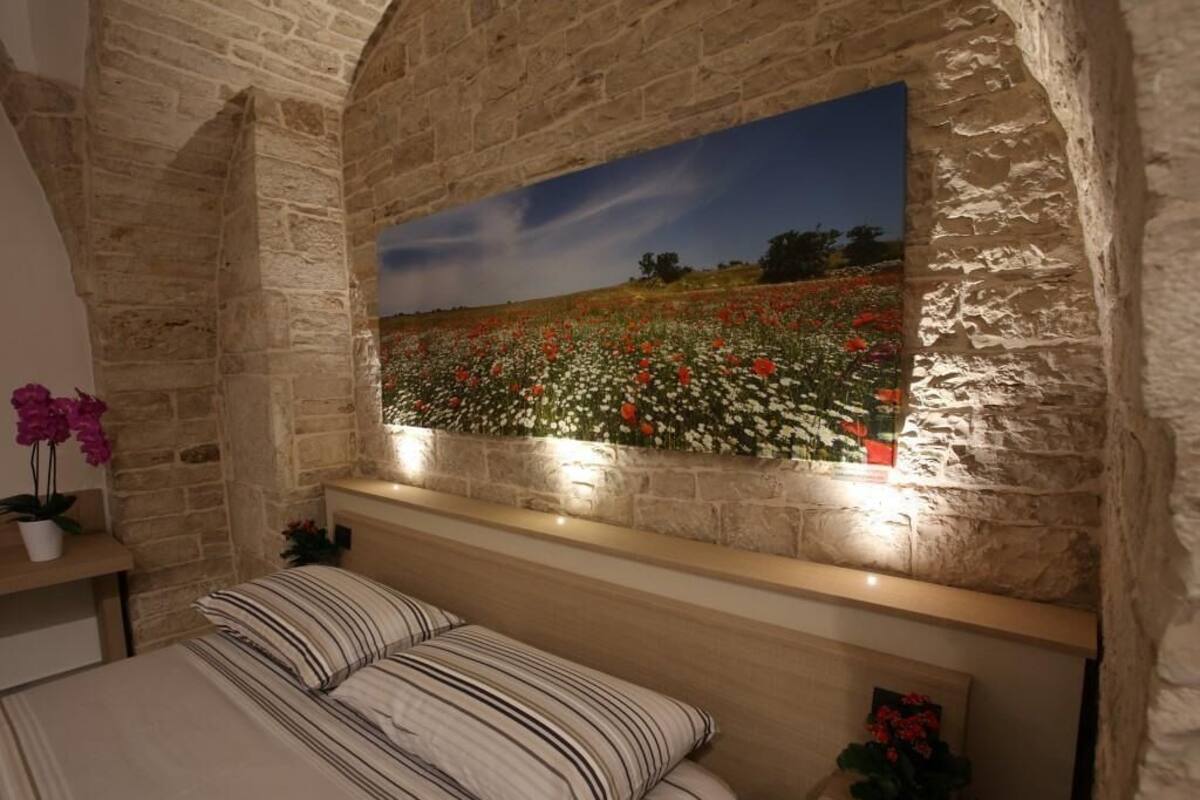 A bedroom with a stone wall, a bed with striped bedding, a nightstand with a potted plant, and a painting of a field.