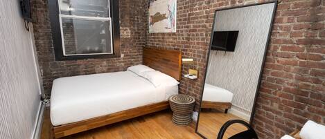 A small, modern room with a wooden bed, a round side table, a mirror, and a brick wall.