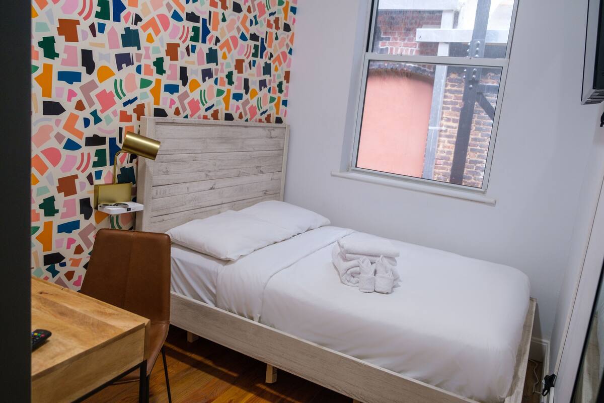 A bedroom with a wooden bed, white bedding, a desk with a chair, a colorful abstract wall, and a window with a view of a brick building.