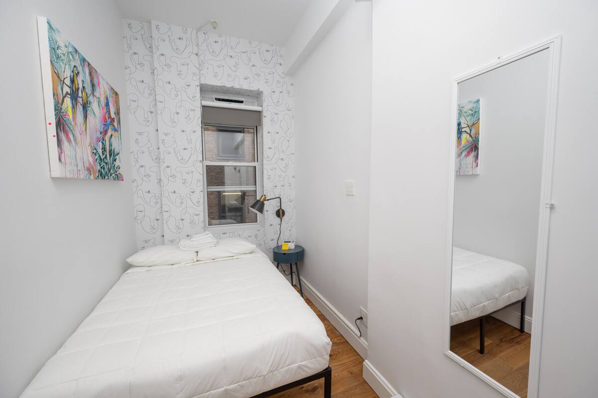 A bedroom with a bed, a window with blinds, a bedside table with a lamp, and a mirror.
