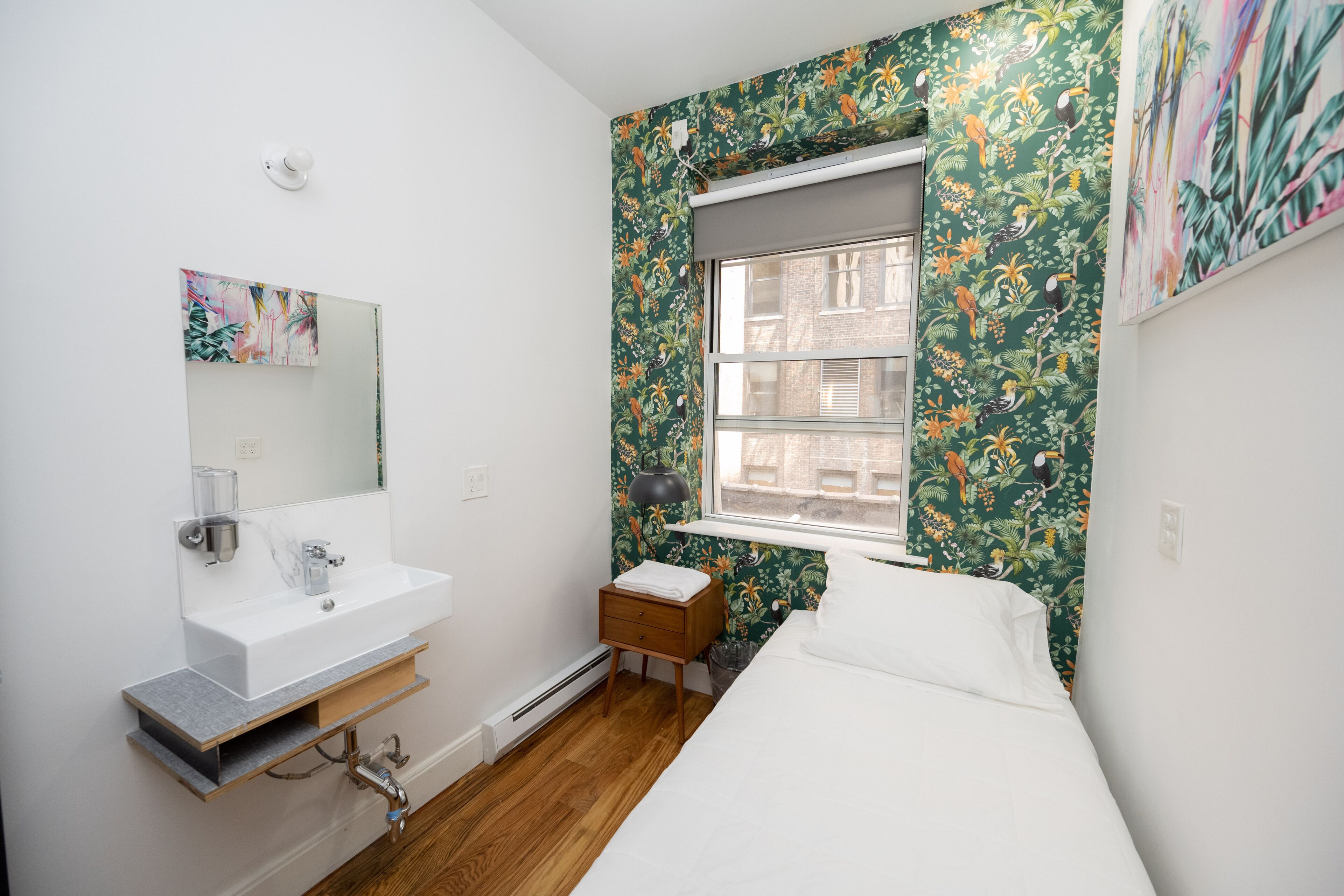 Une petite pièce propre, avec un lavabo blanc, un miroir, un lit et une fenêtre donnant sur des immeubles.