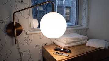 A bedside table with a lamp, a remote control, and a stack of towels.