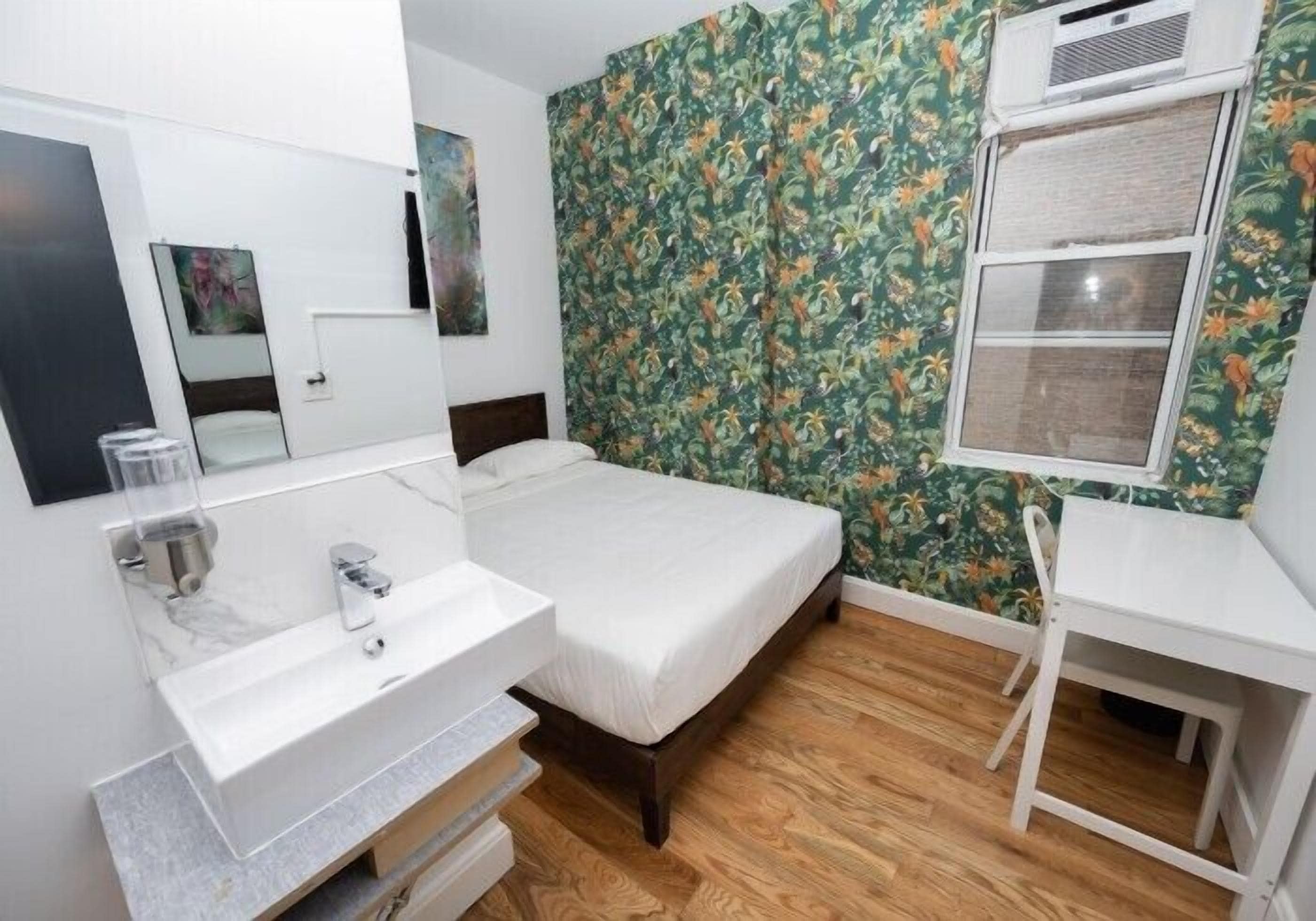 A room with a bed, a white desk, a bathroom with a sink, and a mirror.