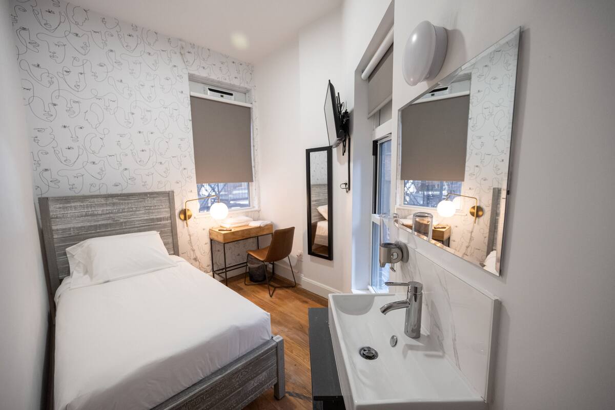 A bedroom with a bed, a desk, and a bathroom with a sink and mirror.