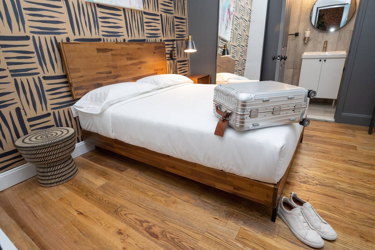 A neatly made bed with a suitcase and shoes on the floor, a round wooden stool, and a wooden headboard.