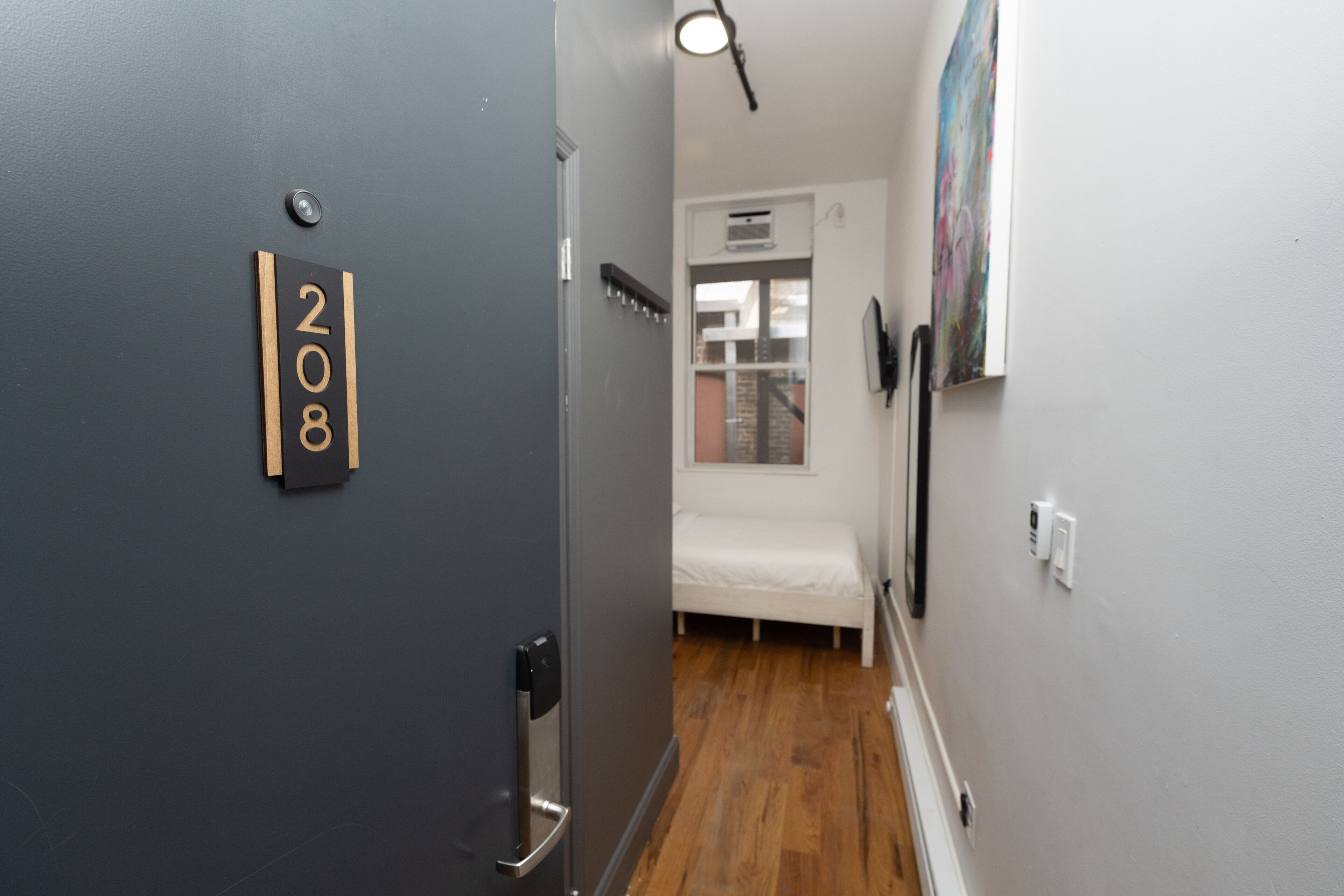 A narrow hallway with a door marked '208', a bed, and a window with an air conditioning unit.